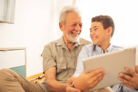 Téléchargez les photos : Garçon et grand-père souriants en vêtements décontractés en utilisant une tablette à la maison pendant le week-end - en image libre de droit