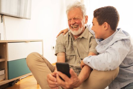 Photo for Smiling grandson in casual clothes teaching laughing grandfather to use smartphone in domestic room at home during weekend - Royalty Free Image