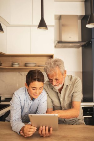 Téléchargez les photos : Heureux adorable garçon enseignant grand-père à utiliser une tablette numérique à la table en bois dans la cuisine à la maison pendant le week-end - en image libre de droit