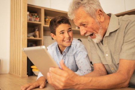 Foto de Sonriente chico lindo viendo abuelo sorprendido usando tableta en la habitación doméstica en casa durante el fin de semana - Imagen libre de derechos