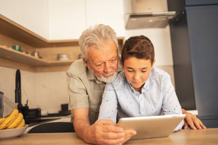 Téléchargez les photos : Heureux homme âgé avec petit-fils en vêtements décontractés regarder le film ensemble sur tablette informatique dans la cuisine à la maison - en image libre de droit