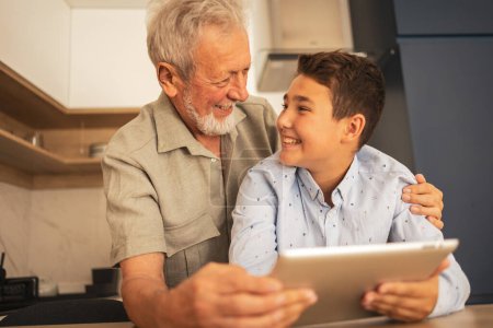 Photo for Smiling grandson and grandfather in casual clothes holding digital tablet while looking at each other in domestic room at home - Royalty Free Image