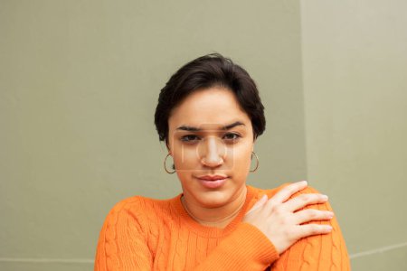 Photo for Portrait of young Latin woman in orange sweater hugging self against beige wall - Royalty Free Image
