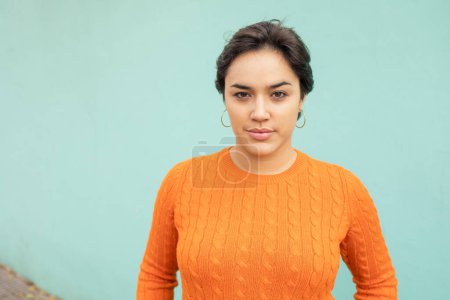 Foto de Retrato de mujer latina joven y confiada en suéter naranja de pie contra la pared turquesa - Imagen libre de derechos