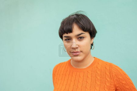Photo for Portrait of stylish young Latin woman in orange sweater with short black hair against turquoise wall - Royalty Free Image