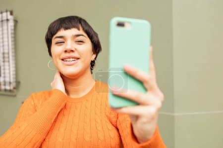 Téléchargez les photos : Joyeux jeune femme latine en pull orange prenant selfie par smartphone contre le mur beige - en image libre de droit