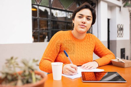 Photo for Thoughtful female Latin freelancer with digital tablet looking away and taking notes in book at sidewalk cafe table - Royalty Free Image