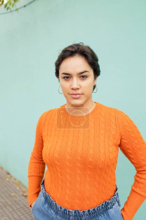 Téléchargez les photos : Confiant jeune femme latine en pull orange debout avec les mains dans les poches contre le mur turquoise en ville - en image libre de droit