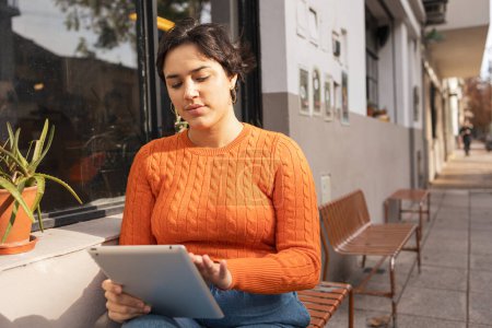 Téléchargez les photos : Jeune femme latine utilisant une tablette numérique alors qu'elle est assise sur un banc en ville pendant la journée - en image libre de droit