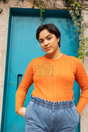 Téléchargez les photos : Portrait de jeune femme latine confiante en pull orange debout avec les mains dans les poches contre porte bleue en ville - en image libre de droit