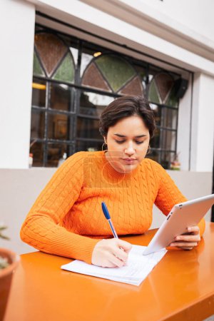 Téléchargez les photos : Jeune indépendante latine prenant des notes dans le livre tout en utilisant une tablette numérique au café trottoir - en image libre de droit