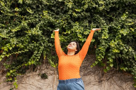 Téléchargez les photos : Jeune femme latine insouciante debout avec les bras levés contre le mur de lierre dans le parc pendant la journée - en image libre de droit