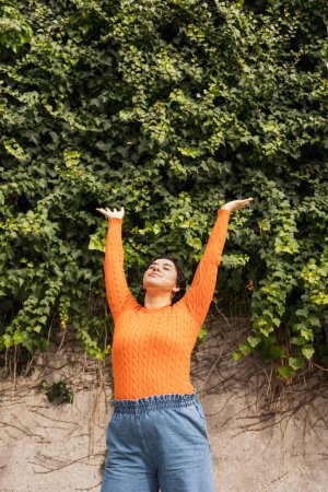 Foto de Relajada joven latina de pie con los brazos levantados contra la pared de hiedra en el parque durante el día - Imagen libre de derechos
