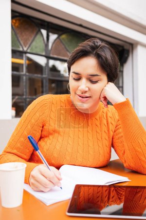 Téléchargez les photos : Confiante jeune femme latine pigiste écriture dans le livre avec tablette numérique sur la table au café trottoir - en image libre de droit