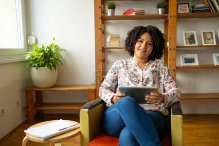 Téléchargez les photos : Portrait de heureuse jeune femme latine avec coiffure afro tenant tablette numérique tout en étant assis sur un fauteuil au bureau à la maison - en image libre de droit