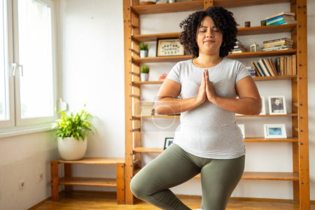 Relaxed young Latin woman in sportswear smiling while practicing tree pose in yoga room at home 