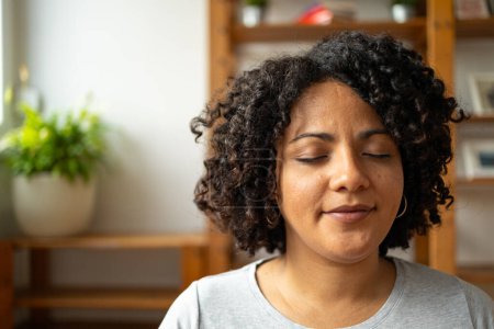 Foto de Mujer latina joven relajada con peinado afro sonriendo mientras medita en la sala de yoga en casa - Imagen libre de derechos
