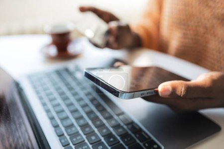 Téléchargez les photos : Jeune pigiste noire méconnaissable tenant smartphone avec ordinateur portable à la table au bureau à la maison - en image libre de droit