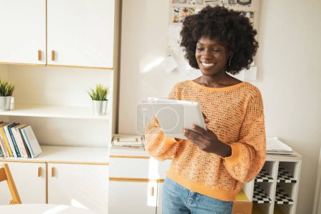 Téléchargez les photos : Heureuse jeune afro-américaine pigiste utilisant une tablette numérique tout en restant au bureau à la maison - en image libre de droit