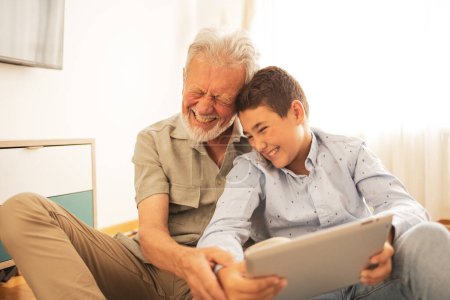 Téléchargez les photos : Joyeux adorable petit-fils et grand-père en vêtements décontractés regarder film drôle sur tablette ensemble à la maison pendant le week-end - en image libre de droit