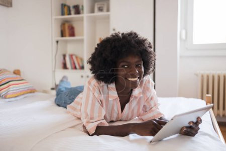 Téléchargez les photos : Pensée heureuse jeune femme noire regardant loin tout en utilisant une tablette numérique dans le lit à la maison - en image libre de droit