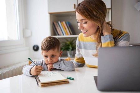 Téléchargez les photos : Souriant jeune mère qui travaille avec un ordinateur portable aider son fils avec des devoirs assis au bureau à la maison - en image libre de droit