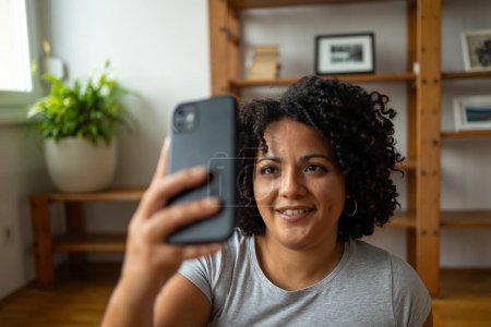 Photo for Content young Latin woman smiling while taking selfie through smartphone in yoga room at home - Royalty Free Image
