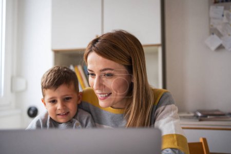 Téléchargez les photos : Séjour souriant à la maison mère aux cheveux blonds travaillant à distance sur ordinateur portable tout en prenant soin de son fils dans l'appartement - en image libre de droit