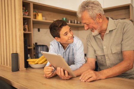 Téléchargez les photos : Souriant grand-père et petit-fils en vêtements décontractés tenant tablette informatique tout en se regardant à la table dans la cuisine à la maison - en image libre de droit