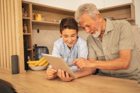 Foto de Nieto sonriente en ropa casual enseñando al abuelo a usar tableta digital en la mesa de madera en la cocina en casa - Imagen libre de derechos