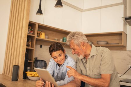 Téléchargez les photos : Heureux grand-père parler avec son petit-fils tout en utilisant une tablette dans la cuisine à la maison pendant le week-end - en image libre de droit