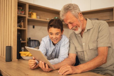 Téléchargez les photos : Souriant homme âgé et petit-fils regarder un film sur tablette numérique ensemble à la table en bois dans la cuisine à la maison pendant le week-end - en image libre de droit