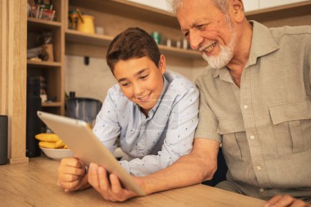 Téléchargez les photos : Souriant grand-père et petit-fils en vêtements décontractés regarder film sur tablette informatique à table dans l'appartement pendant le week-end - en image libre de droit