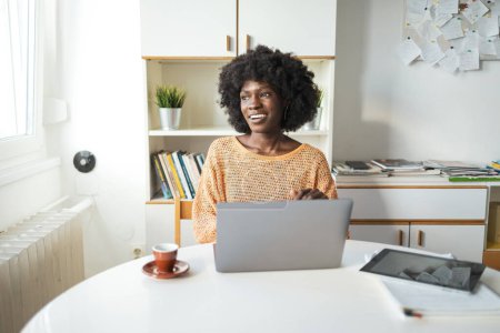 Photo for Thoughtful young black female freelancer looking away while sitting with laptop and digital tablet at table in home office - Royalty Free Image