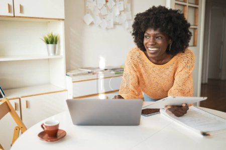 Photo for Happy thoughtful young black female freelancer looking away while using laptop and digital tablet at table in home office - Royalty Free Image