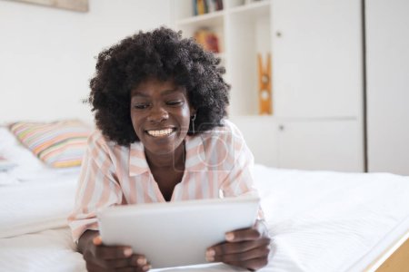 Foto de Feliz joven afroamericana con peinado afro usando tableta digital mientras está acostado en la cama en casa - Imagen libre de derechos