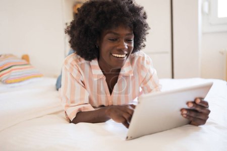 Téléchargez les photos : Heureuse jeune femme noire avec une coiffure afro à l'aide d'un ordinateur tablette tout en étant couchée au lit à la maison - en image libre de droit