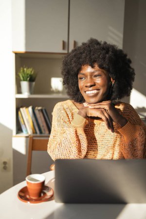 Foto de Feliz mujer negra joven reflexiva mirando hacia otro lado mientras descansa la barbilla en las manos mientras está sentado con el ordenador portátil en la mesa en la oficina en el hogar - Imagen libre de derechos