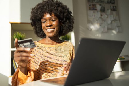 Photo for Smiling young African American woman using credit card and laptop for online shopping at home - Royalty Free Image