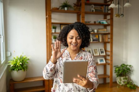 Photo for Happy young Latin female freelancer waving at digital tablet during video call at home office - Royalty Free Image