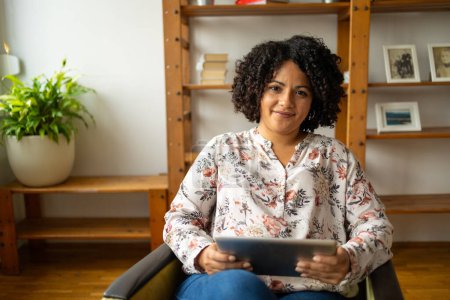 Photo for Portrait of happy young Latin woman holding digital tablet while sitting on armchair at home office - Royalty Free Image