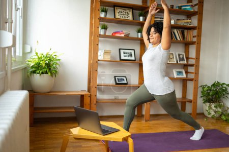 Téléchargez les photos : Heureuse jeune femme latine en vêtements de sport pratiquant dans la posture guerrière dans la salle de yoga pendant les cours en ligne sur ordinateur portable à la maison - en image libre de droit