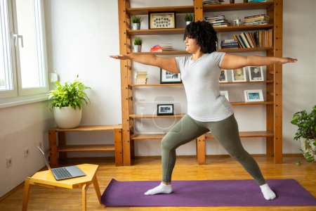 Photo for Full body of young Latin woman in sportswear practicing yoga in warrior pose on yoga mat during online classes at home - Royalty Free Image