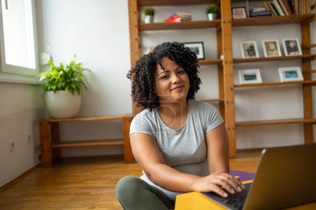 Foto de Retrato de una joven latina sonriente con peinado afro usando portátil mientras está sentada en el suelo en la sala de yoga en casa - Imagen libre de derechos