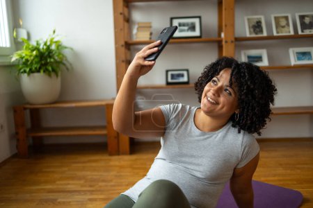 Téléchargez les photos : Joyeux jeune femme latine avec afro coiffure prendre selfie par smartphone tout en étant assis sur tapis de yoga à la maison - en image libre de droit
