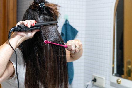 Téléchargez les photos : Gros plan de jeune femme redressant les cheveux avec un fer à repasser en céramique dans sa salle de bain à la maison. Concept de soins capillaires. Photo de haute qualité - en image libre de droit