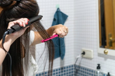 Téléchargez les photos : Gros plan de jeune femme redressant les cheveux avec un fer à repasser en céramique dans sa salle de bain à la maison. Concept de soins capillaires. Photo de haute qualité - en image libre de droit