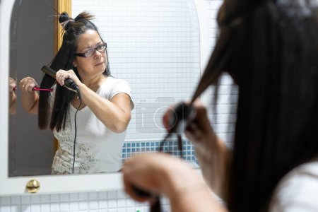 Téléchargez les photos : Femme redressant ses cheveux avec un fer à repasser en céramique se regardant dans le miroir de salle de bain à la maison. Concept de soins capillaires. Photo de haute qualité - en image libre de droit