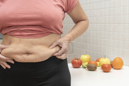 Frau mittleren Alters zeigt überschüssiges Bauchfett und im Hintergrund verschiedenes Obst und Gemüse. Konzept der Gewichtsabnahme durch gesunde Ernährung. Hochwertiges Foto