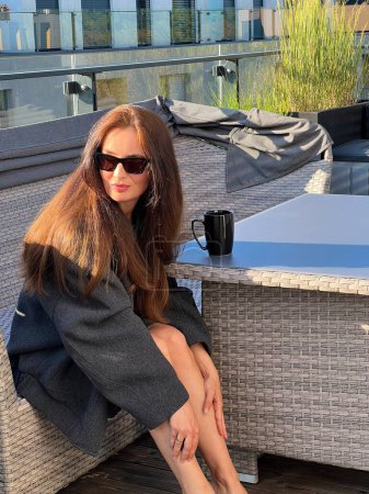 On a sunny day, a young adult woman wearing black sunglasses and a grey oversized bomber jacket sits on the terrace next to wicker furniture, with a black cup resting on the table. Slow life concept.